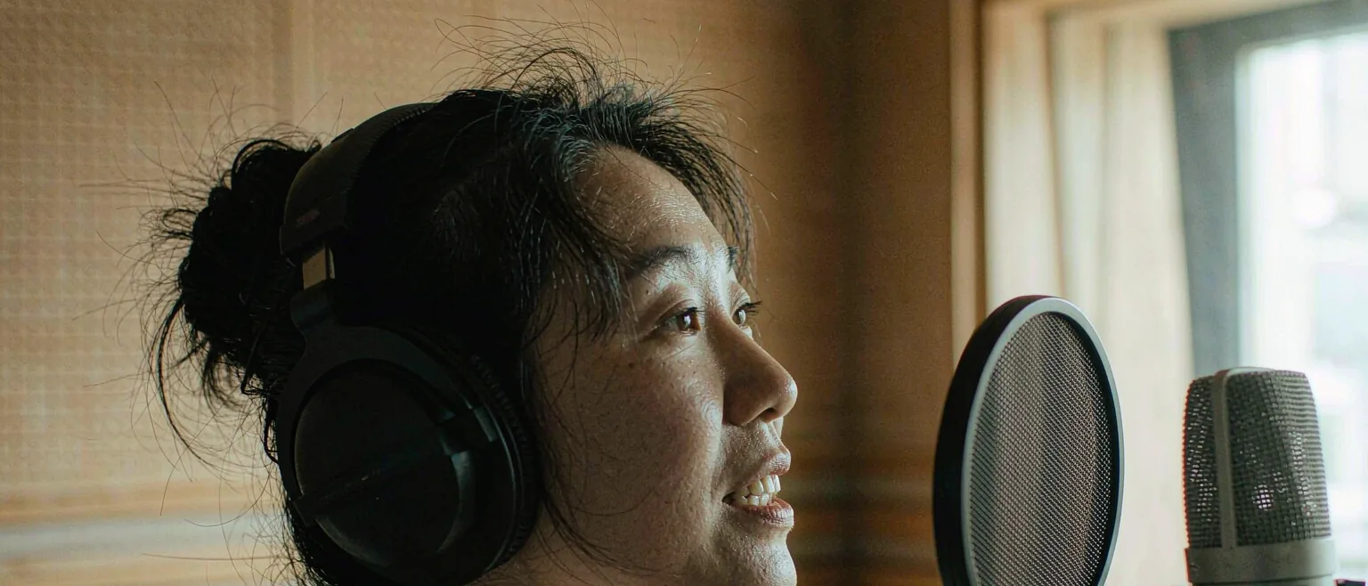 中国女性在录音棚内对着专业麦克风说话,暖色木质吸音墙与午后侧光