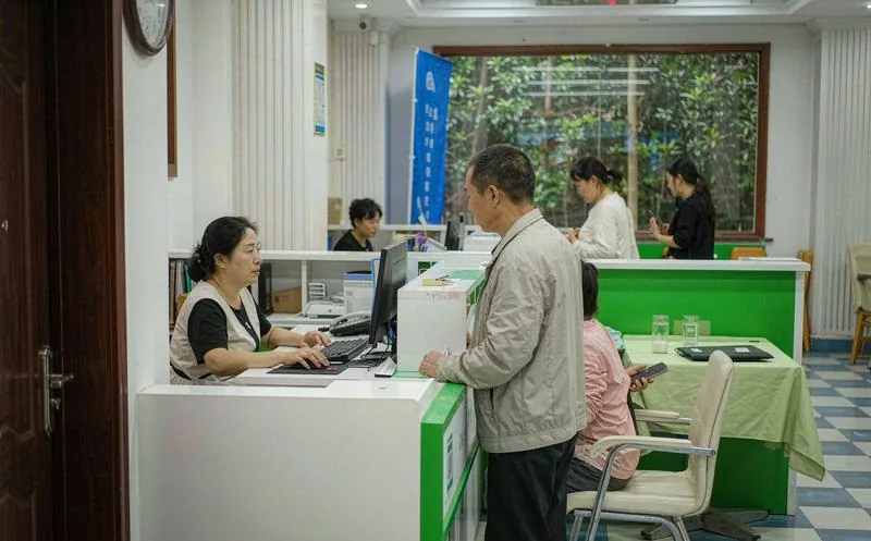 换热站机房内一名穿深蓝色工装的中国男性技术人员正弯腰检查板式换热器管路接口,背景可见多台循环水泵、压力表和保温管道,机房灯光偏冷白,地面有反光,景深中隐约可见墙上运行看板
