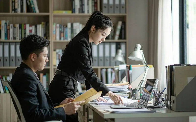 中国代理公司的会议室内,两名穿职业装的中文员工在堆满案卷的桌前翻阅文件并核对墙上进度看板,自然光,专业办公氛围,写实摄影