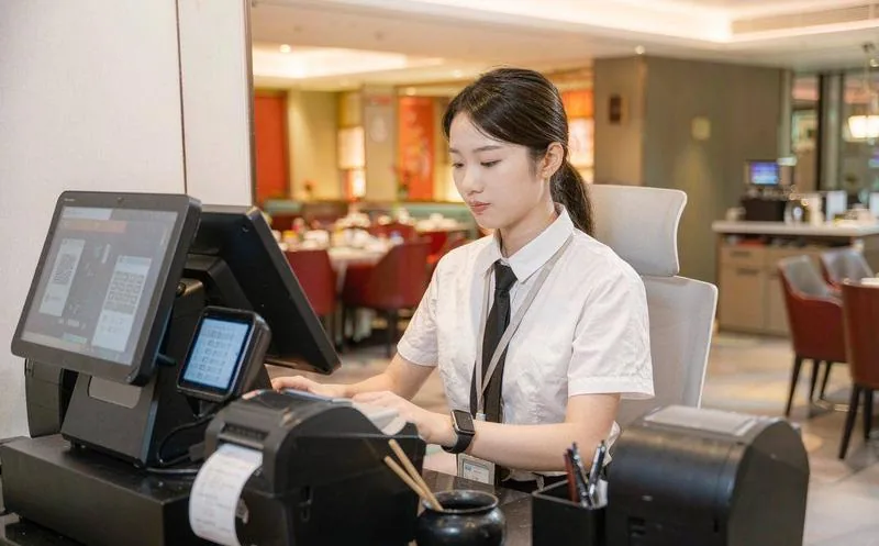 中国连锁餐饮门店里,一名店长在中午高峰时段查看前台收银与后厨出餐屏,背景是忙碌的点餐区