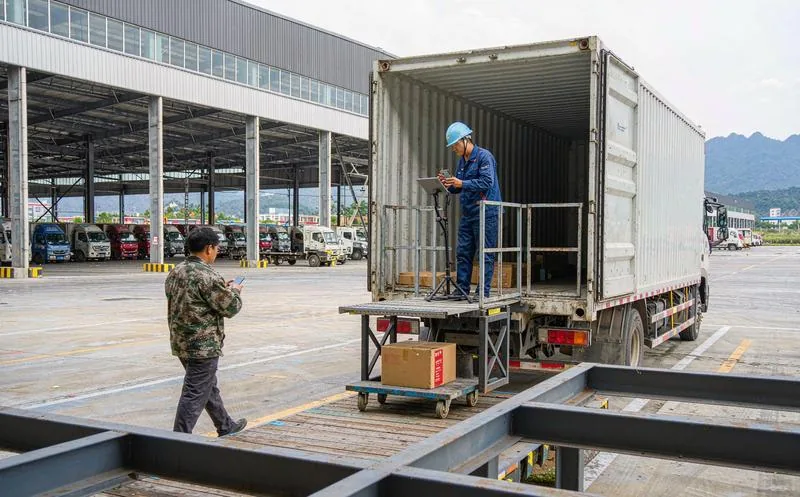 中国物流园区月台前,戴蓝色安全帽的员工站在货车尾板用平板电脑扫描登记纸箱,背景停车场上卡车整齐排列,写实摄影